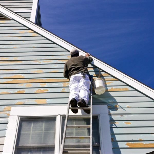 Painter scraping and prepping peeling siding and trim near the roofline before repainting